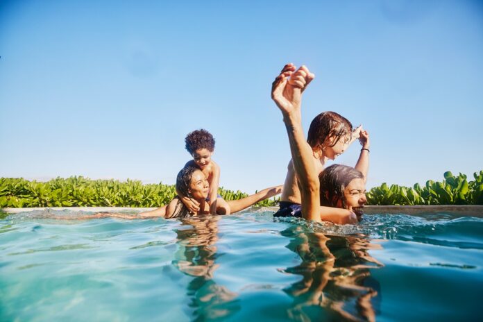 Wide shot of smiling sons riding on mothers backs while playing in pool at tropical resort